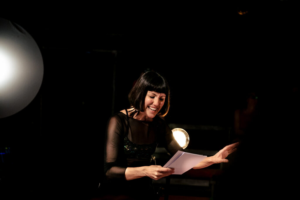 A promotional image for the show Selene. A woman with dark hair and closes smiles while reading a script, one hand raised, backed by two circular lights