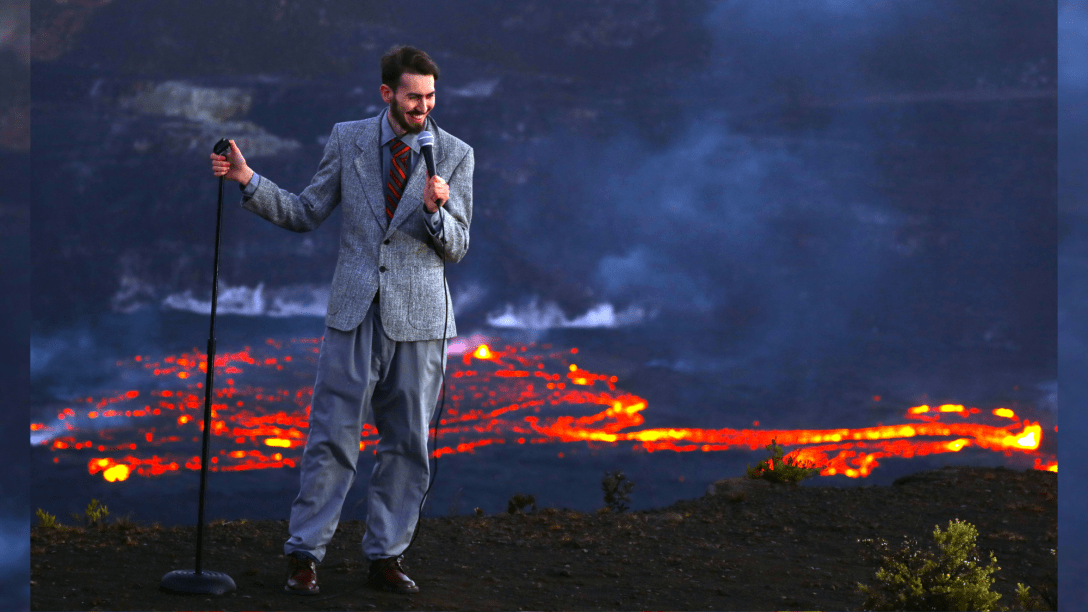 A talk white bearded man in a grey suit holding a microphone in one hand and the microphone stand in the other, stand in from of an active lava field.