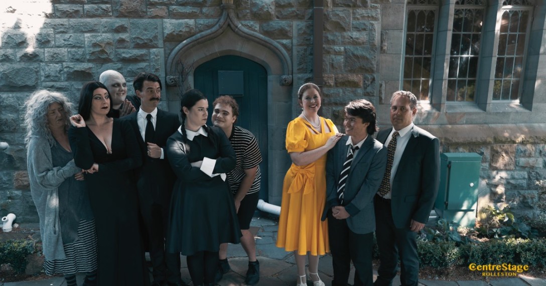 The cast of the musical The Addams Family in costume, starnding in character in front of a stone wall