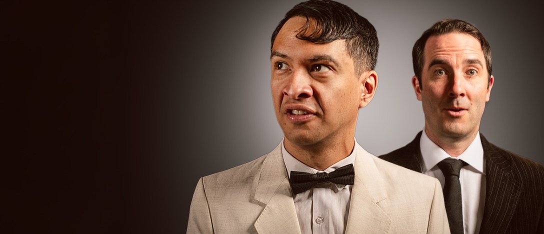 A shoulder-up photo of two actors with startled expressions, both wearing suits