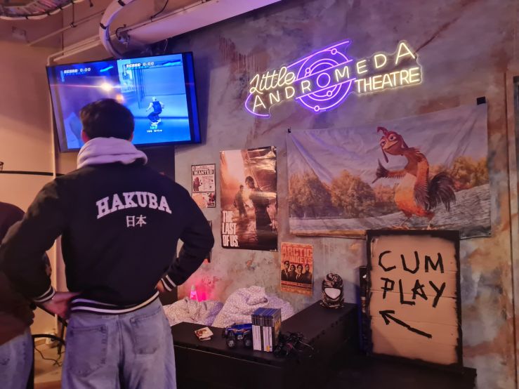 A recreation of a guy's bedroom in the corner of the Little Andromeda foyer. Some men with their backs to the camera play a skateboarding game on a playstation.