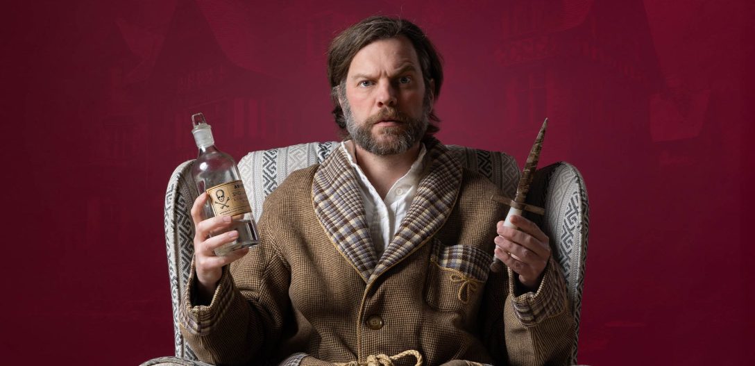 a confused looking man in a tweed smoking jacket looks at the camera, holding a bottle of poison in one hand and a dagger in the other.