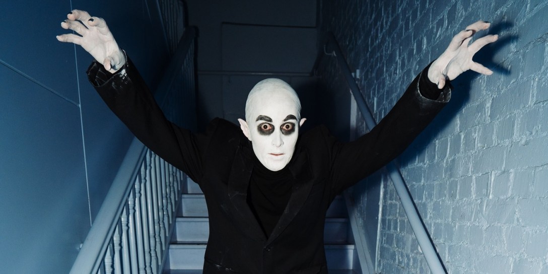 A bald vampire with dark eyes, whist skin and block clothes stands in a stairwell, raising his arms and leaning towards the camera