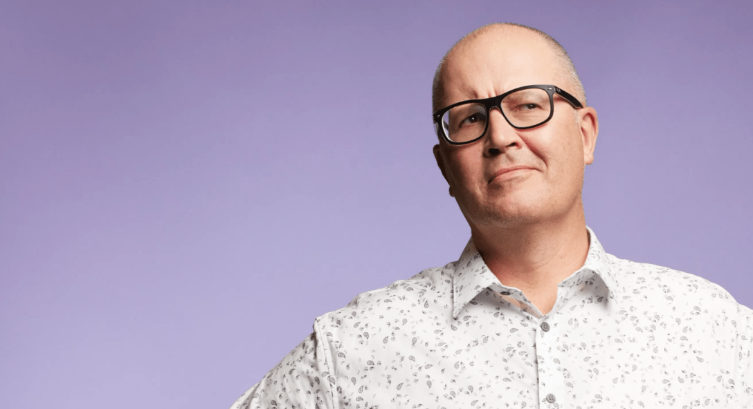 A bald white man with glasses in a white buttong up shirt - comedian Paul Ego - against a lilac backdrop