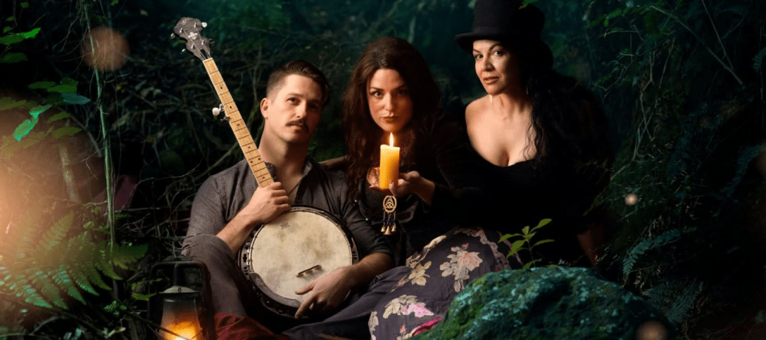 A white man holding a banjo, a white woman holding a candle and a Māori woman in a top hat are seated ono the ground, looking at the camera, surrounded by ferns and greenery.