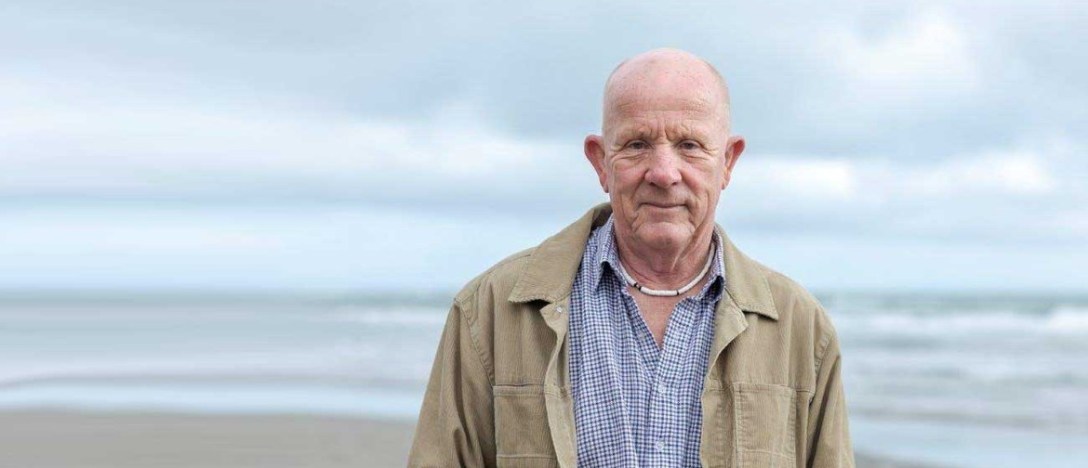 A photograph of writer Joe Bennet standing on a beach with this back to the water.